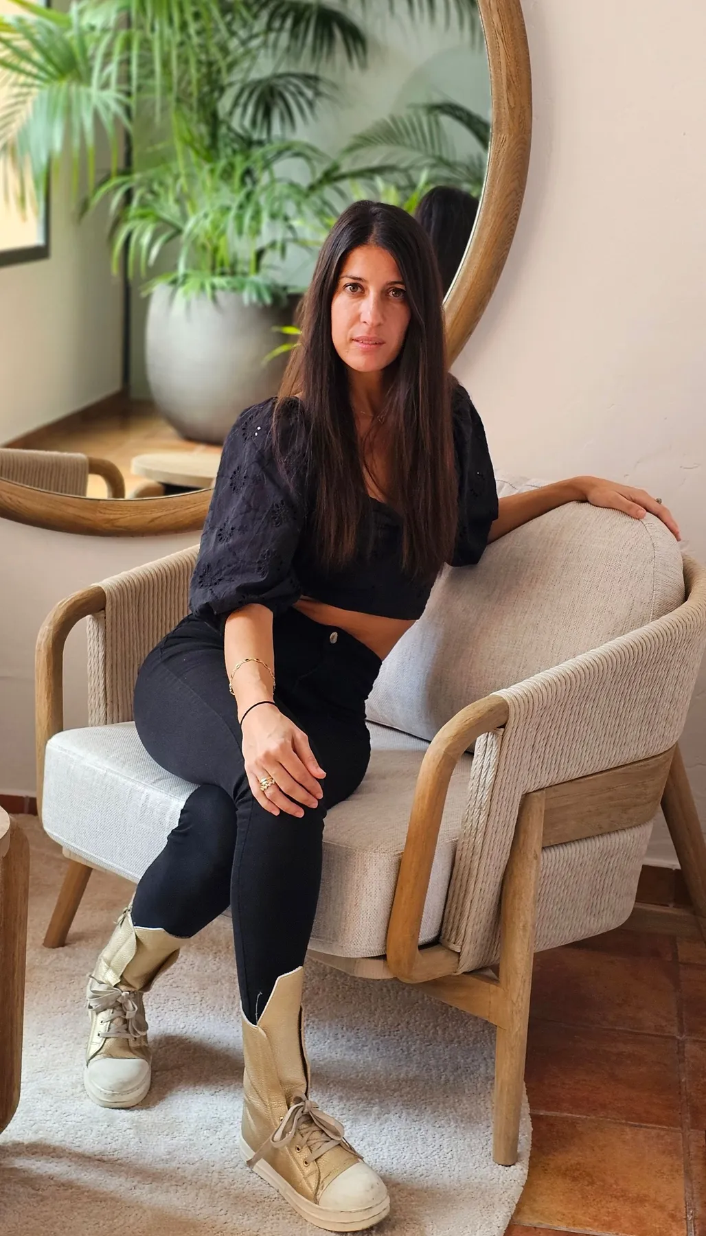 Laura sitting in an armchair beside a round mirror and plants